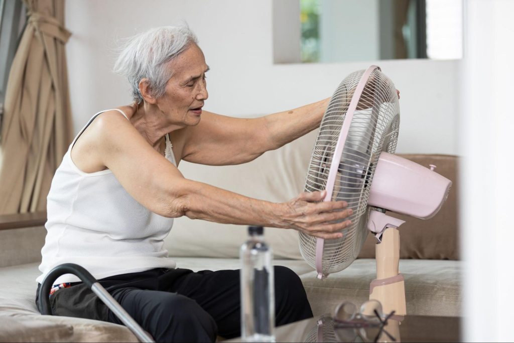 Uma idosa na frente de um ventilador se refrescando, perto uma garrafa de água