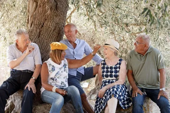 Um grupo de idosos, homens e mulheres, sentados ao redor de uma árvore rindo e conversando.