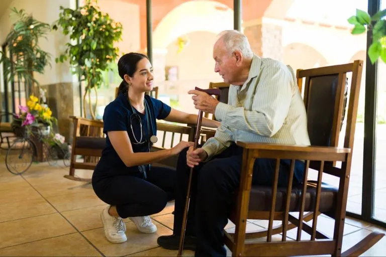 Um idoso sentado em uma cadeira de balanço acompanhado de uma médica abaixada conversando.