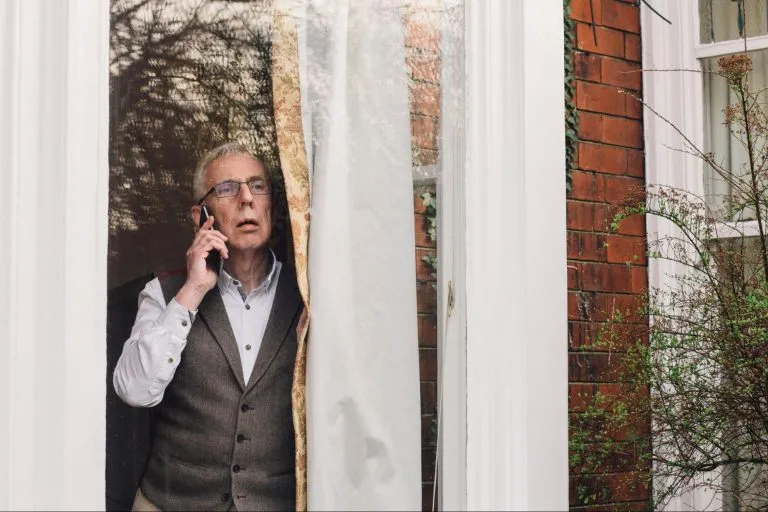 Um idoso em uma janela olhando para fora com um telefone na mão