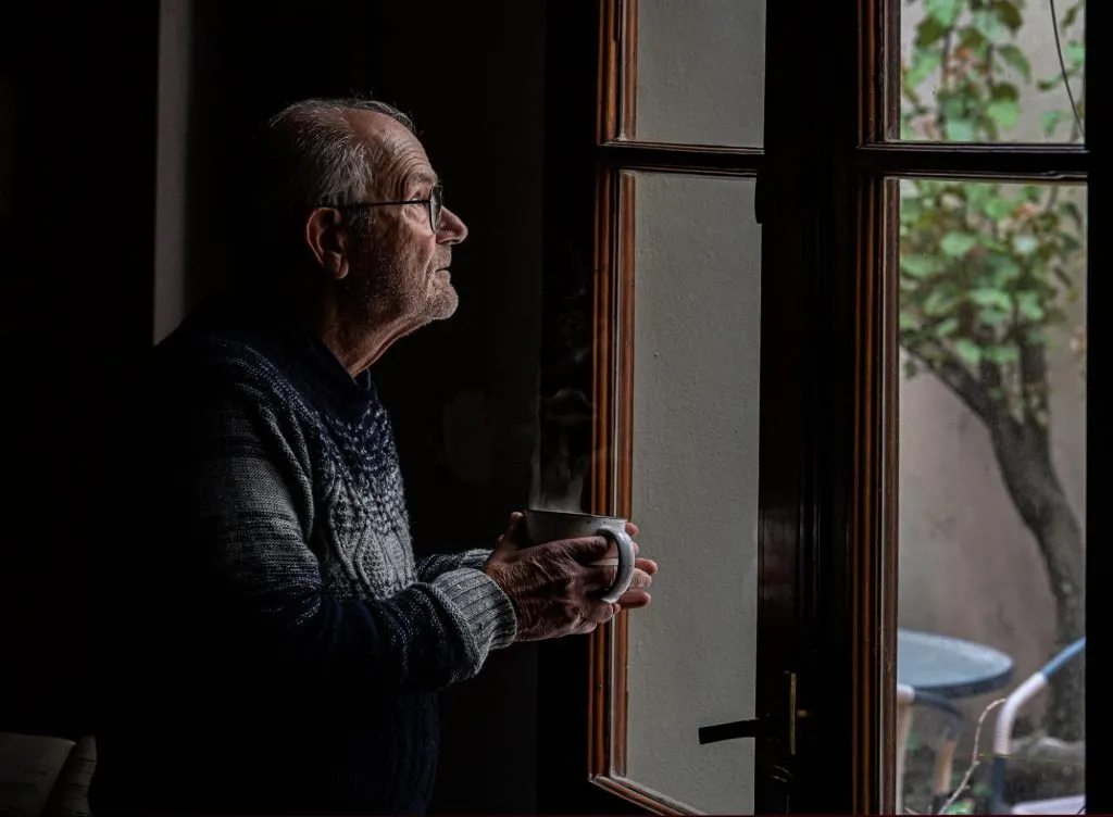 Um idoso segurando uma caneca olhando através de uma janela.