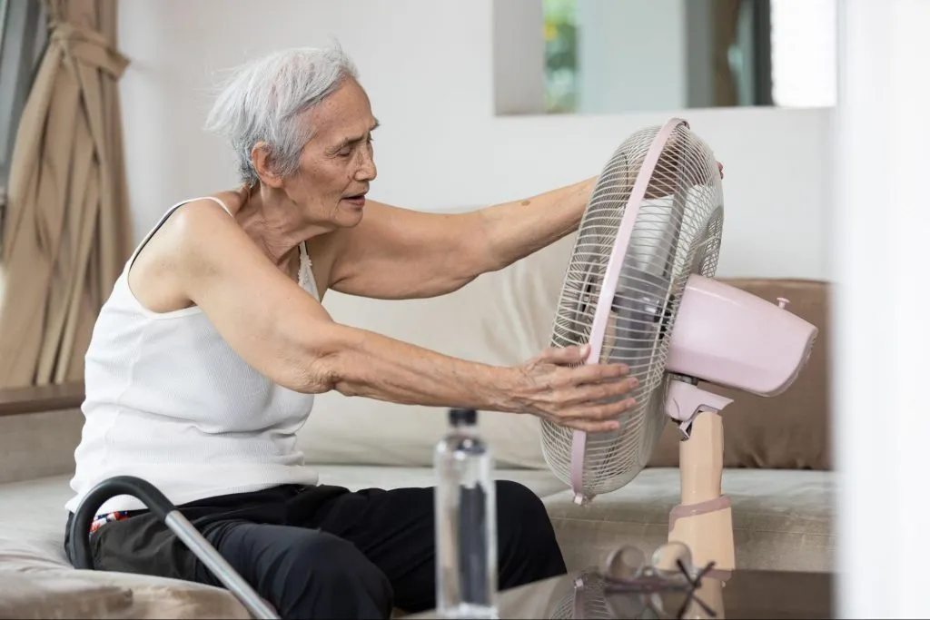 Uma idosa na frente de um ventilador se refrescando, perto uma garrafa de água